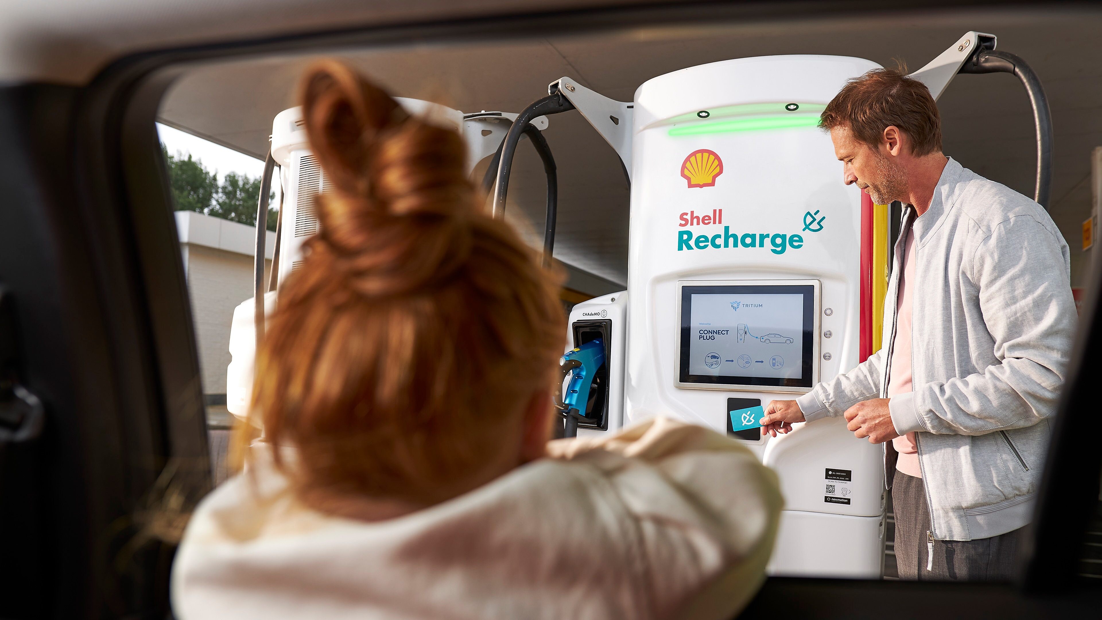 A man using a Shell Recharge unit while a girl watches from inside a car